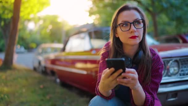 ヘッドフォン付きの女性は 赤いヴィンテージカーの隣で曲がり 夏の日に咲く庭や緑の通りでスマートフォンを使用しています ファッショナブルなトレンド — ストック動画