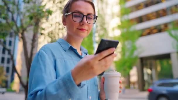 戴眼镜的白人女人在城市里走来走去 用智能手机 喝咖啡 摩天大楼在后面 工作日 忙碌生活概念 — 图库视频影像