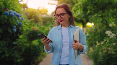 Dizüstü bilgisayarı ve akıllı telefonu olan mutlu bir kadın çiçek açan bahçede ya da işten sonra yeşil sokakta yürüyor, ağır çekimde..