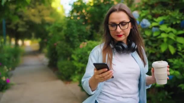 夏の日に咲く庭や緑の通りを歩いている彼女の手のコーヒーとスマートフォンでメガネとヘッドフォンで幸せな女性 — ストック動画
