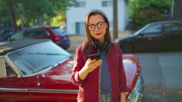 ヘッドフォン付きの女性は赤いヴィンテージカーの近くで屋外に立っており 夏の日に咲く庭や緑の通りでスマートフォンを使用しています ファッショナブルなトレンド — ストック動画