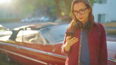 Kulaklık takan kadın dışarıda, kırmızı vintage arabanın yanında duruyor ve yaz günü bahçesinde ya da yeşil sokakta akıllı telefon kullanıyor. Moda eğilimi. 