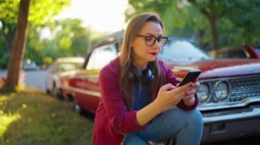 Kulaklıklı bir kadın kırmızı vintage bir arabanın yanına çömelmiş ve yaz günü yeşil bir sokakta akıllı telefon kullanıyor. Moda eğilimi. 