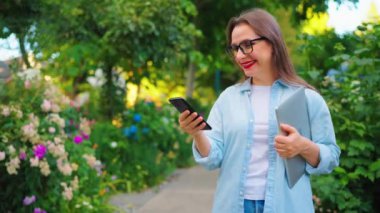 Dizüstü bilgisayarı ve elinde akıllı telefonu olan mutlu bir kadın çiçek açan bahçede ya da işten sonra yeşil sokakta duruyor, ağır çekim..