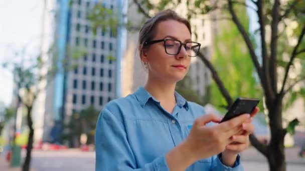 戴眼镜的白人女人在城市里走来走去 用智能手机 摩天大楼在后面 工作日 忙碌生活概念 — 图库视频影像