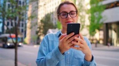 Gözlüklü beyaz kadın şehrin ortasında duruyor ve akıllı telefon kullanıyor, ağır çekimde. Arka planda gökdelenler ve arabalar var. İletişim, iş günü, yoğun yaşam konsepti.