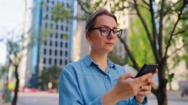 Gözlüklü beyaz kadın şehirde dolaşıyor ve akıllı telefon kullanıyor, ağır çekimde. Arka planda gökdelenler var. İletişim, iş günü, yoğun yaşam konsepti.
