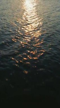 Dikey video. Gün batımında deniz veya okyanusun mavi yüzeyinin havadan görünüşü. Güneş dalgalara yansıyor ve parıldıyor altın denizi aydınlatıyor.