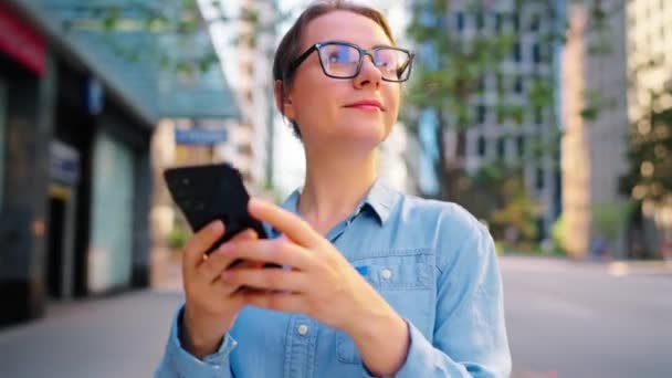 眼鏡をかけた白人女性は 街の真ん中に立ち スマートフォンを使用しています 超高層ビルやクルマが背景に立っている コミュニケーション 仕事の日 忙しい人生の概念 — ストック動画