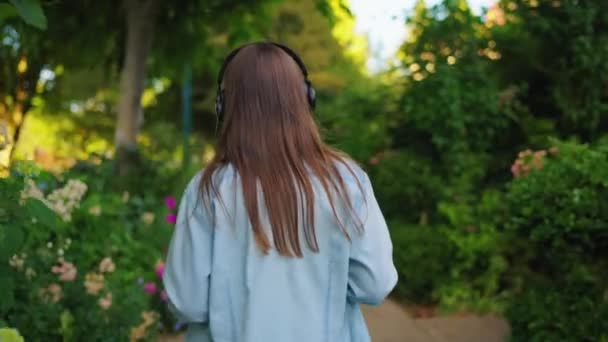 戴着眼镜 头戴耳机 手里拿着咖啡和智能手机的快乐女性正走在盛开的花园或绿色的街道上 动作缓慢 — 图库视频影像