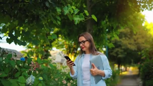 ラップトップとスマートフォンのハッピーな女性は 仕事の日の後に咲く庭や緑の通りを歩いています — ストック動画