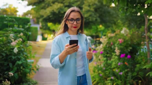 快乐的女人 手里拿着笔记本电脑和智能手机 下班后漫步在繁华的花园或绿色的街道上 动作缓慢 — 图库视频影像