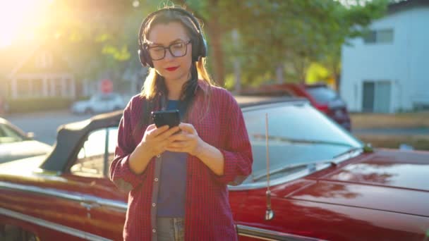 Kobieta Słuchawkami Stoi Zewnątrz Pobliżu Czerwonego Samochodu Vintage Pomocą Smartfona — Wideo stockowe
