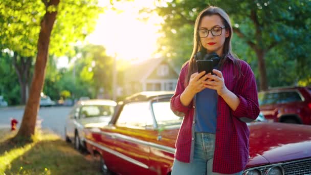 ヘッドフォン付きの女性は赤いヴィンテージカーの近くで屋外に立っており 夏の日に咲く庭や緑の通りでスマートフォンを使用しています ファッショナブルなトレンド — ストック動画