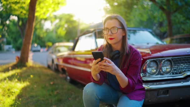 Женщина Наушниках Приседает Рядом Красной Винтажной Машиной Использует Смартфон Цветущем — стоковое видео