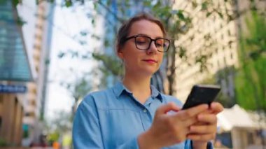 Gözlüklü beyaz kadın şehirde dolaşıyor ve akıllı telefon kullanıyor. Arka planda gökdelenler var. İletişim, iş günü, yoğun yaşam konsepti.