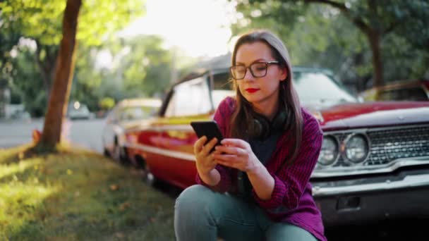 Женщина Наушниках Приседает Рядом Красной Винтажной Машиной Использует Смартфон Цветущем — стоковое видео