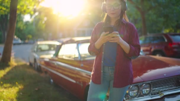 ヘッドフォン付きの女性は赤いヴィンテージカーの近くで屋外に立っており 夏の日に咲く庭や緑の通りでスマートフォンを使用しています ファッショナブルな傾向 スローモーション — ストック動画