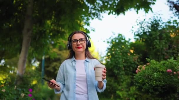 Šťastná Žena Brýlích Sluchátkách Kávou Smartphonem Rukou Kráčející Kvetoucí Zahradou — Stock video