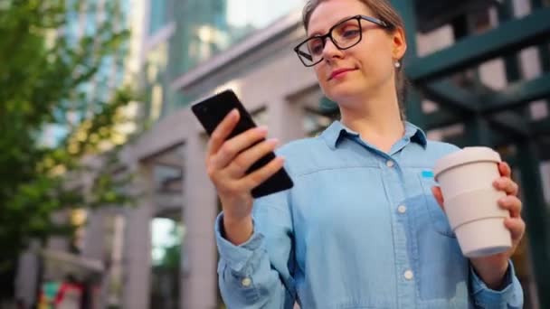 戴眼镜的白人妇女站在街上 用智能手机 喝咖啡 轨道射击 摩天大楼在后面 工作日 忙碌生活概念 — 图库视频影像