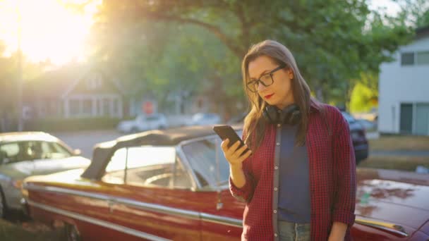 女性は赤いヴィンテージカーの近くで屋外に立っており 夏の日に咲く庭や緑の通りでスマートフォンを使用しています ファッショナブルなトレンド — ストック動画