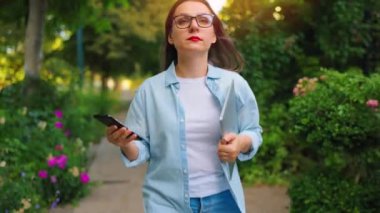 Dizüstü bilgisayarı ve akıllı telefonu olan mutlu bir kadın çiçek açan bahçede ya da işten sonra yeşil sokakta yürüyor..