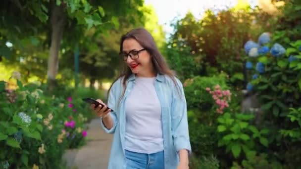 快乐的微笑的女人 手里拿着智能手机 下班后漫步在繁华的花园或绿色的街道上 — 图库视频影像