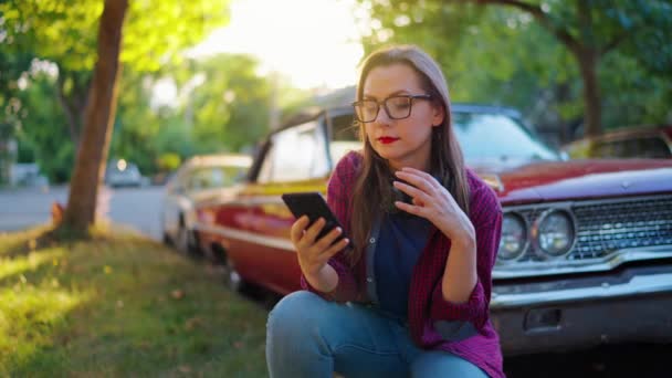 ヘッドフォン付きの女性は 赤いヴィンテージカーの隣で曲がり 夏の日に咲く庭や緑の通りでスマートフォンを使用して ゆっくり動きます ファッショナブルなトレンド — ストック動画