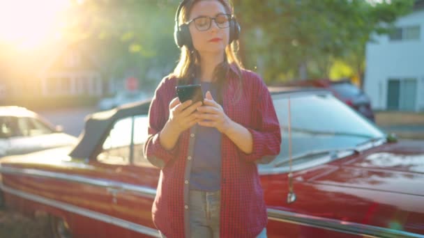 ヘッドフォン付きの女性は赤いヴィンテージカーの近くで屋外に立っており 夏の日に咲く庭や緑の通りでスマートフォンを使用しています ファッショナブルな傾向 スローモーション — ストック動画