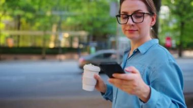 Gözlüklü beyaz bir kadın şehirde geziyor, akıllı telefon kullanıyor ve kahve içiyor. Şehir arka planda. İletişim, iş günü, yoğun yaşam konsepti.