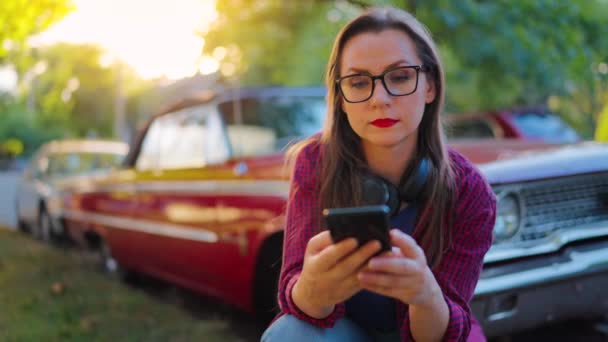 ヘッドフォンが付いている女性は 赤いヴィンテージ車の隣で曲がり 夏の日に緑の通りでスマートフォンを使用しています ファッショナブルなトレンド — ストック動画