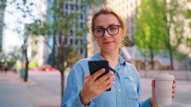 Gözlüklü beyaz kadın sokakta duruyor, akıllı telefon kullanıyor ve kahve içiyor, ağır çekimde. Arka planda gökdelenler var. İletişim, iş günü, yoğun yaşam konsepti.