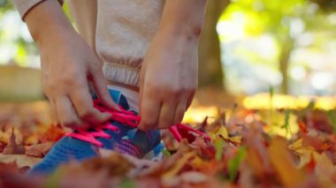 Gün batımında koşarken ya da yürürken ayakkabı bağı bağlayan bir kadın. Sonbahar sarı yaprakları. Yavaş çekim.