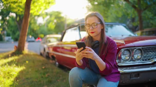 ヘッドフォンが付いている女性は 赤いヴィンテージ車の隣で曲がり 夏の日に緑の通りでスマートフォンを使用しています ファッショナブルなトレンド — ストック動画