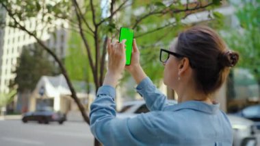 Kadın sokakta dikiliyor ve yeşil ekran, yörünge görüntülü akıllı telefon ile fotoğraf çekiyor. Yavaş çekim.