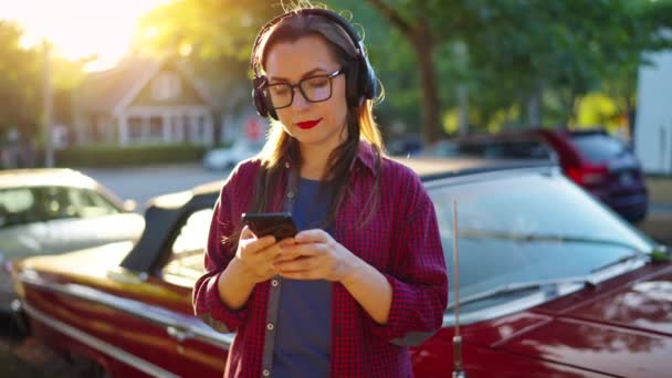 ヘッドフォン付きの女性は赤いヴィンテージカーの近くで屋外に立っており 夏の日にグリーンストリートでスマートフォンを使用しています ファッショナブルな傾向 スローモーション — ストック動画