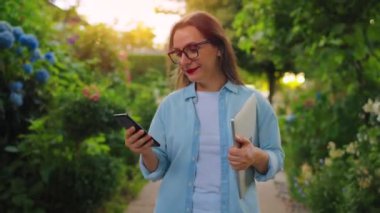Dizüstü bilgisayarı ve akıllı telefonu olan mutlu bir kadın çiçek açan bahçede ya da işten sonra yeşil sokakta yürüyor..