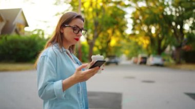 Happy woman with the smartphone and coffee in her hands walking through the neighborhood in summer evening.