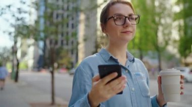 Gözlüklü beyaz bir kadın şehirde geziyor, akıllı telefon kullanıyor ve kahve içiyor. Şehir arka planda. İletişim, iş günü, yoğun yaşam konsepti.
