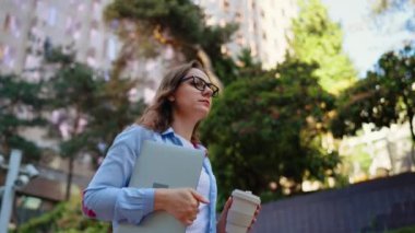 Carefree business woman walking on the street with laptop and coffee in hands after work day in the office. Slow motion