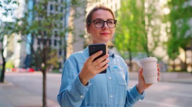 Gözlüklü beyaz kadın sokakta duruyor, akıllı telefon kullanıyor ve kahve içiyor. Yörünge atışı. Arka planda gökdelenler var. İletişim, iş günü, yoğun yaşam konsepti.