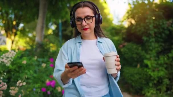 Gözlüklü Kulaklıklı Mutlu Kadın Elinde Kahve Akıllı Telefonuyla Çiçek Açan — Stok video