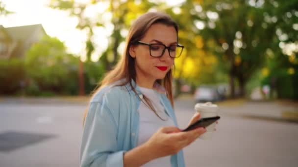 夏の夕方に近所を歩いている彼女の手のスマートフォンとコーヒーの幸せな女性 スローモーション — ストック動画