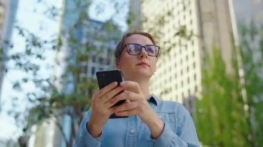 Gözlüklü beyaz kadın şehrin caddesinde dikiliyor ve akıllı telefon kullanıyor. Arka planda gökdelenler var. İletişim, iş günü, yoğun yaşam konsepti.