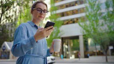 Beyaz gözlüklü kadın cadde boyunca yürüyor, akıllı telefon kullanıyor ve kahve içiyor. Yörünge atışı. Arka planda gökdelenler var. İletişim, iş günü, yoğun yaşam konsepti.