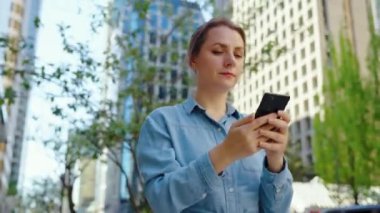 Beyaz bir kadın şehirde dolanıyor ve akıllı telefon kullanıyor. Arka planda gökdelenler var. İletişim, iş günü, yoğun yaşam konsepti. 