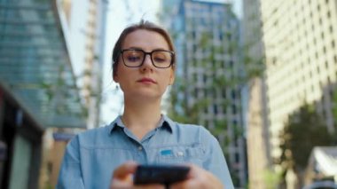 Gözlüklü beyaz kadın şehirde dolanıyor ve akıllı telefon kullanıyor. Arka planda gökdelenler var. İletişim, iş günü, yoğun yaşam konsepti. Yavaş çekim