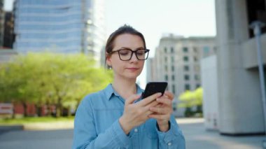 Gözlüklü beyaz kadın şehirde dolanıyor ve akıllı telefon kullanıyor. Arka planda gökdelenler var. İletişim, iş günü, yoğun yaşam konsepti.