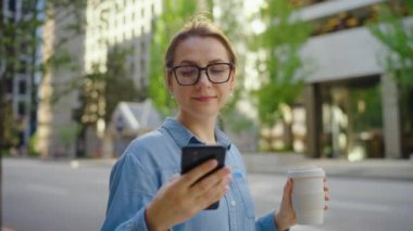 Gözlüklü beyaz kadın sokakta duruyor, akıllı telefon kullanıyor ve kahve içiyor. Yörünge atışı. Arka planda gökdelenler var. İletişim, iş günü, yoğun yaşam konsepti.