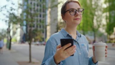 Beyaz gözlüklü kadın cadde boyunca yürüyor, akıllı telefon kullanıyor ve kahve içiyor. Yörünge atışı. Arka planda gökdelenler var. İletişim, iş günü, yoğun yaşam konsepti.